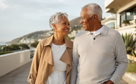 An older couple walks outdoors, smiling at each other. They are casually dressed and appear happy, with buildings, greenery, and hills in the background under a partly cloudy sky.