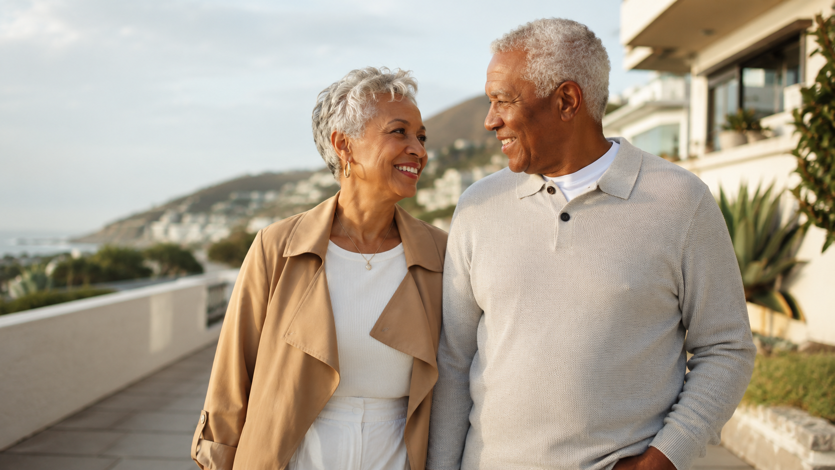 An older couple walks outdoors, smiling at each other. They are casually dressed and appear happy, with buildings, greenery, and hills in the background under a partly cloudy sky.