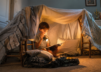 A young child sits inside a cozy blanket fort, reading a book by flashlight. Pillows and soft blankets make the fort inviting, while warm light creates a magical, nighttime atmosphere in a softly lit room.