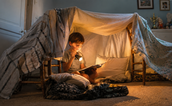 A young child sits inside a cozy blanket fort, reading a book by flashlight. Pillows and soft blankets make the fort inviting, while warm light creates a magical, nighttime atmosphere in a softly lit room.