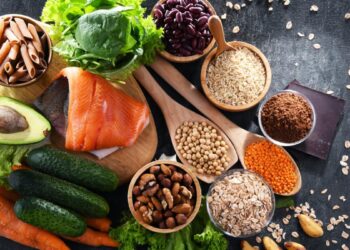 A variety of healthy foods on a dark surface, including salmon, spinach, avocado, pasta, beans, rice, nuts, seeds, oats, red lentils, carrots, cucumbers, lettuce, and a piece of dark chocolate—perfect for eating for longevity.