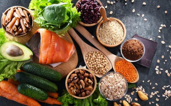 A variety of healthy foods on a dark surface, including salmon, spinach, avocado, pasta, beans, rice, nuts, seeds, oats, red lentils, carrots, cucumbers, lettuce, and a piece of dark chocolate—perfect for eating for longevity.