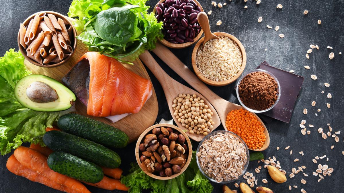A variety of healthy foods on a dark surface, including salmon, spinach, avocado, pasta, beans, rice, nuts, seeds, oats, red lentils, carrots, cucumbers, lettuce, and a piece of dark chocolate—perfect for eating for longevity.