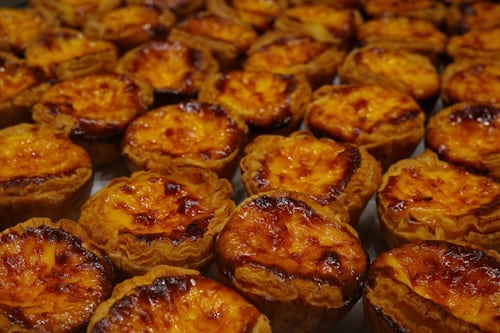 Rows of golden-brown Portuguese custard tarts (pastéis de nata) with caramelized tops and flaky pastry crusts, arranged closely together.