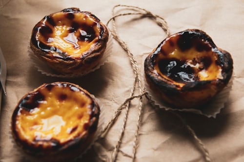 Three Portuguese custard tarts (pastéis de nata) with golden, caramelized tops sit on brown parchment paper, next to a piece of rustic twine.