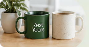 Two mugs sit on a wooden table: one is green with “Zest Years” printed on it, the other is plain cream. A leafy plant in a white pot sits behind them—perfect for a lifestyle site for older adults like About ZestYears.
