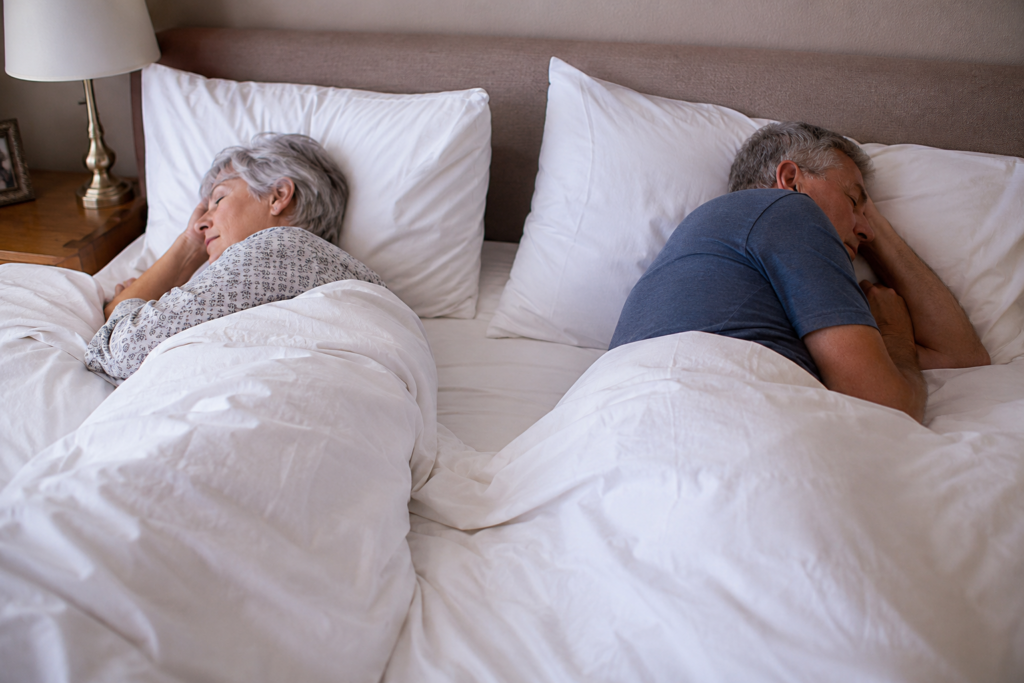 Two older adults, a woman and a man, sleep on opposite sides of a bed under white blankets, facing away from each other. The softly lit room hints at the quiet distance that sometimes leads couples to consider separate bedrooms in marriage.