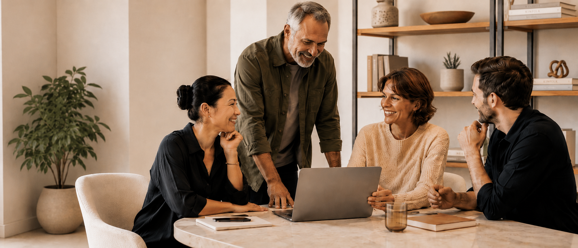 Four people sit around a table in a modern office, smiling and talking. One stands while others sit with a laptop, notebooks, and a glass of water on the table. Explore careers at ZestYears in this welcoming workspace with shelves and greenery in the background.