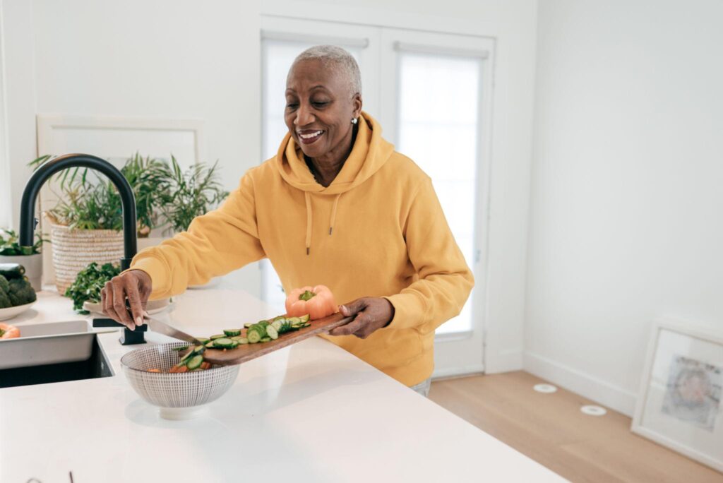 An older woman wearing a yellow hoodie smiles while adding chopped vegetables from a wooden board into a colander, embracing food as medicine in her bright, modern kitchen.