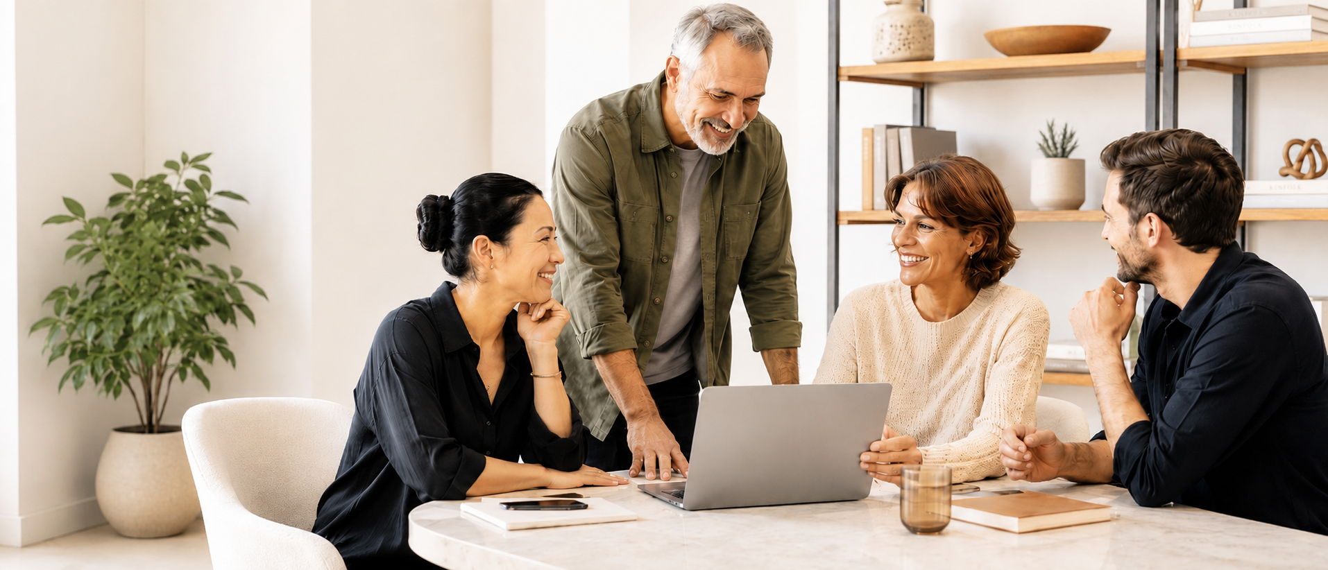 Four adults sit and stand around a table, smiling and talking while looking at a laptop. The setting is a bright, modern office with shelves and a plant in the background—an inviting atmosphere that perfectly captures the collaborative spirit of Careers at ZestYears.