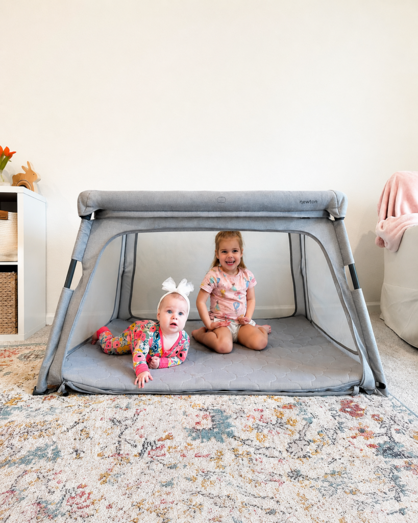 Two young children, a baby and a toddler, are inside a gray playpen on a patterned rug. The baby is lying on her stomach, and the toddler is sitting, both smiling at the camera in a bright, cozy room.