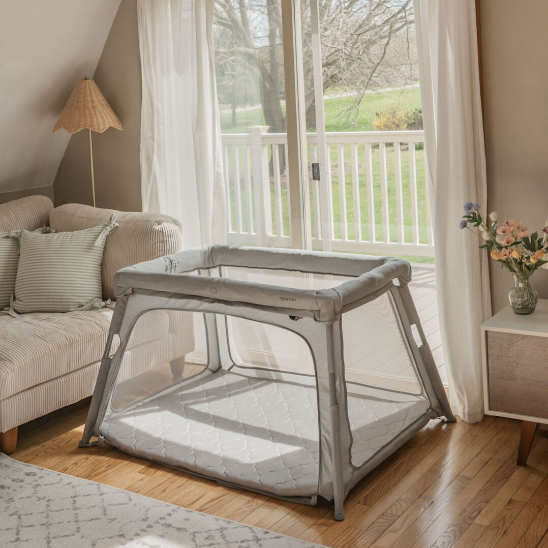 A gray playpen sits on a wooden floor in a cozy living room, next to a beige sofa, a side table with flowers, and large glass doors that open to a sunny deck and green lawn outside.