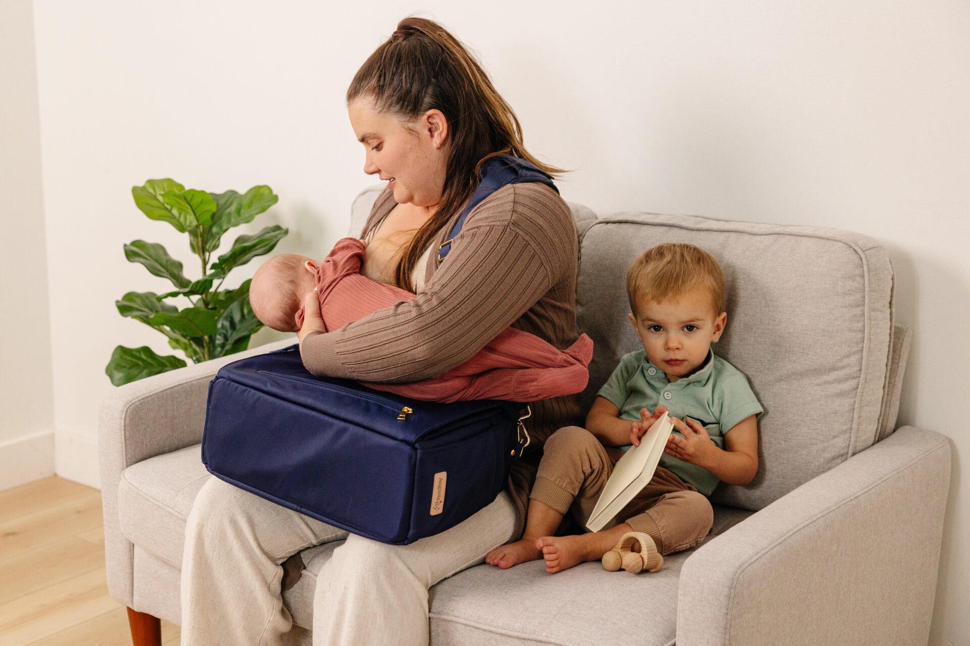 A woman breastfeeds a baby while sitting on a couch. A young child sits beside her holding an open book. There is a blue bag on the womans lap and a green plant in the corner.