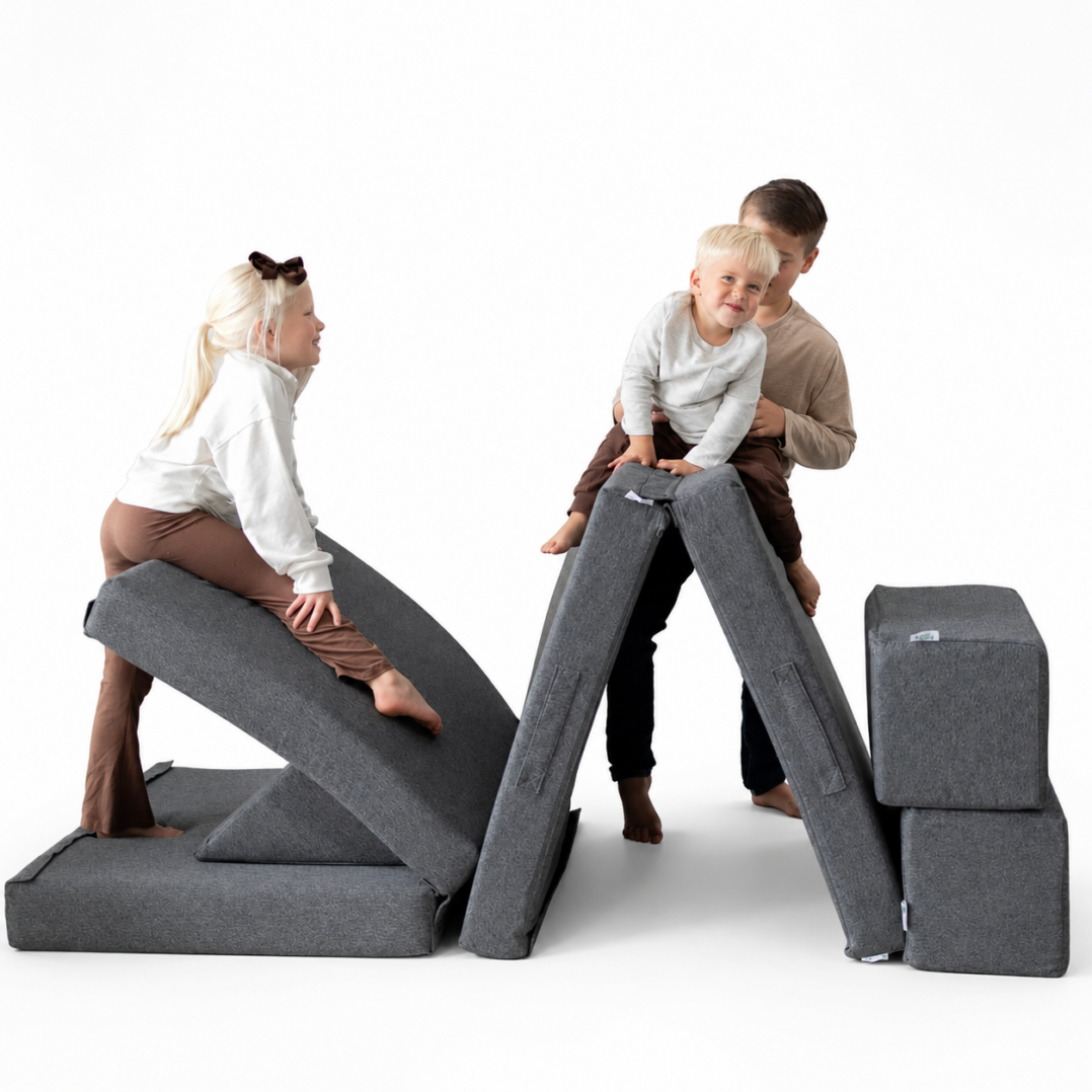 Three children play with modular grey foam cushions, arranging them into creative shapes for climbing and sitting, against a plain white background.