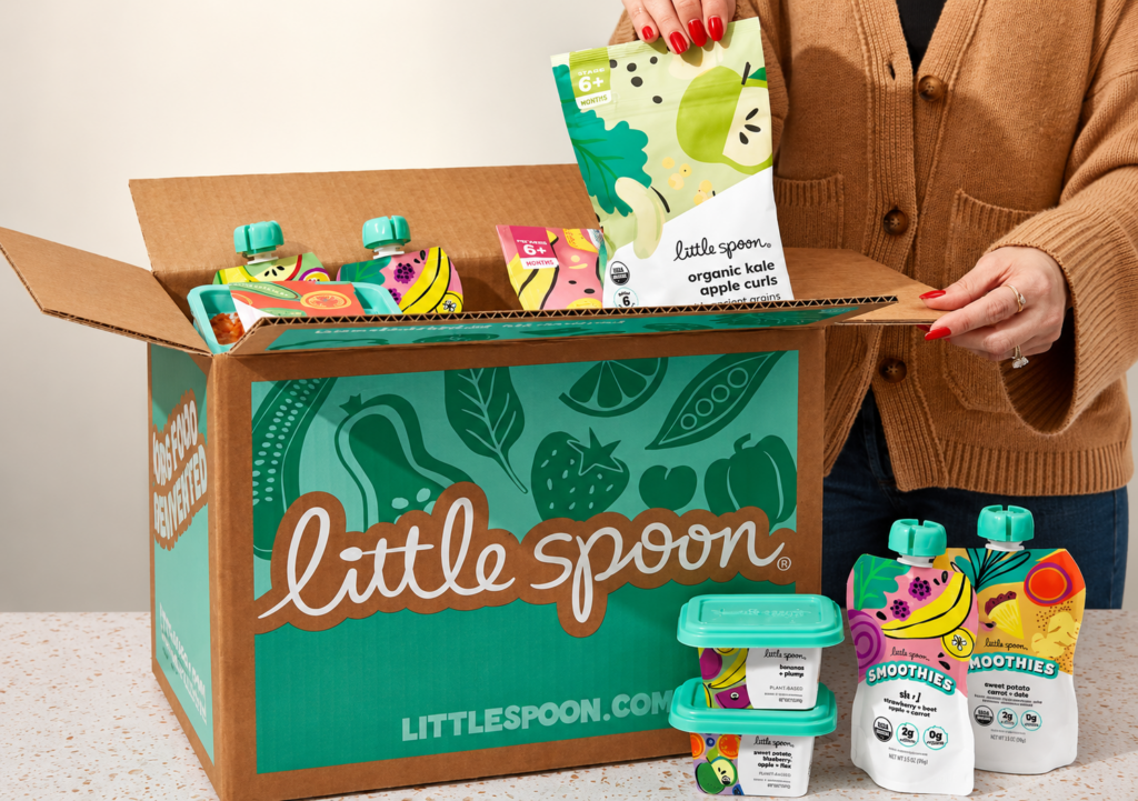 A person holds a pouch of Little Spoon organic kale apple curls above an open box filled with various Little Spoon baby food pouches and containers. Several pouches and containers are displayed on the counter nearby.