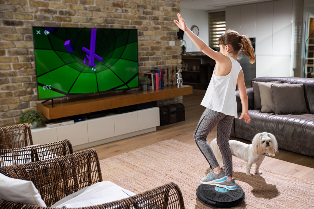 A girl balances on a circular game controller in a living room, playing an interactive game displayed on a TV. A small white dog stands beside her on the rug, and the room has modern decor with a brick accent wall.