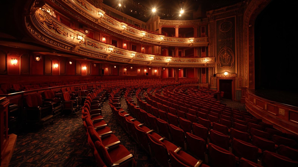 A grand, ornate theater with rows of red velvet seats, gold-trimmed balconies, and warm lighting, facing an empty stage. The ambience is elegant and classic, with intricate architectural details visible.