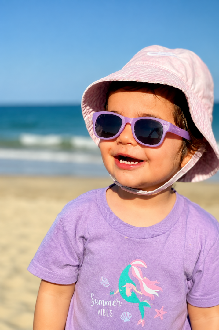A young child wearing a sun hat, purple sunglasses, and a lavender Summer Vibes t-shirt smiles while standing on a sandy beach with the ocean and blue sky in the background.