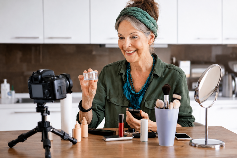 An older woman, acting as a senior product tester, sits at a kitchen table recording a video. She holds up a cosmetic jar and smiles, with makeup products, brushes, and a mirror arranged on the table in front of her.
