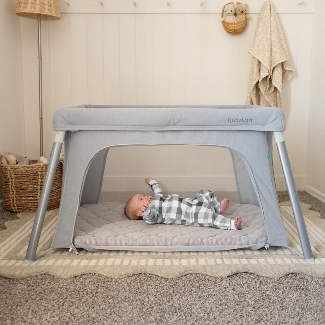 A baby in a gray checkered onesie lies on their back inside a light gray playpen with metal legs, in a cozy, softly lit room with a basket and hanging clothes in the background.