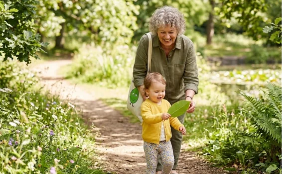 An older woman and a toddler walk along a sunny forest path. The child holds a large green leaf, smiling, while the woman follows behind, also smiling. Lush greenery and wildflowers surround them.
