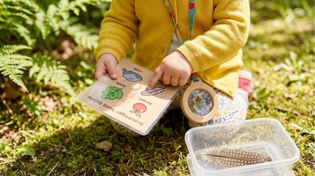 A child in a yellow sweater points to a scavenger hunt guide outdoors, with a clear magnifier and a box containing a feather nearby, surrounded by green plants and grass.