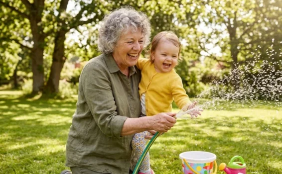 An older woman and a young child laugh together while playing with a garden hose, spraying water outside on a sunny day. A toy bucket and watering can sit on the grass nearby.