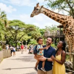 A smiling family with a small child stands near a giraffe at a zoo on a sunny day. Trees and other visitors are visible in the background.