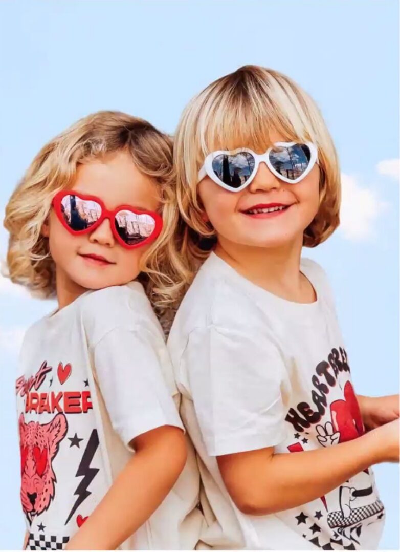Two young children with blonde hair wear heart-shaped sunglasses and matching graphic t-shirts, smiling and posing playfully against a blue sky background.