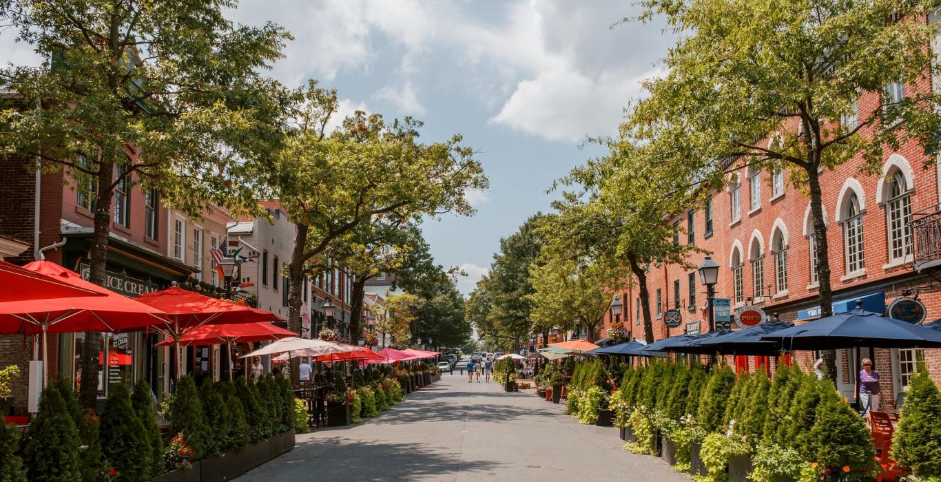 A lively street scene with outdoor dining, red and blue umbrellas, green plants, and brick buildings lining both sides under a partly cloudy sky.