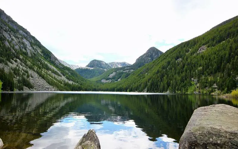 A calm mountain lake reflects green forested slopes and rocky peaks under a cloudy sky, with boulders in the foreground.