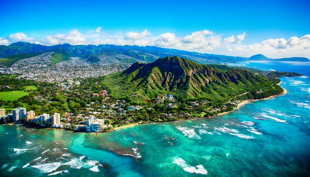 Aerial view of a lush green volcanic mountain near turquoise ocean waters, with a city and coastline in the background under a blue sky with scattered clouds.