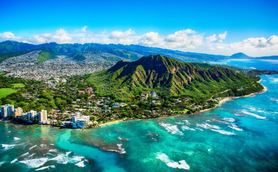 Aerial view of a lush green volcanic mountain near turquoise ocean waters, with a city and coastline in the background under a blue sky with scattered clouds.