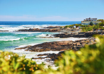 Rocky coastline with waves crashing, lush green bushes in the foreground, and a large house overlooking the ocean on a sunny day.