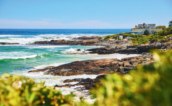 Rocky coastline with waves crashing, lush green bushes in the foreground, and a large house overlooking the ocean on a sunny day.