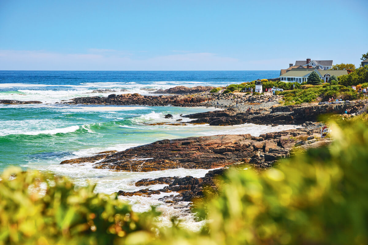 Rocky coastline with waves crashing, lush green bushes in the foreground, and a large house overlooking the ocean on a sunny day.
