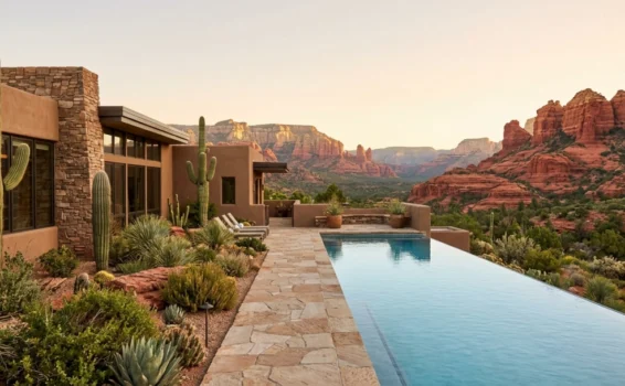 A modern house with stone walls and cacti sits beside an infinity pool, overlooking red rock formations and desert landscape at sunset.