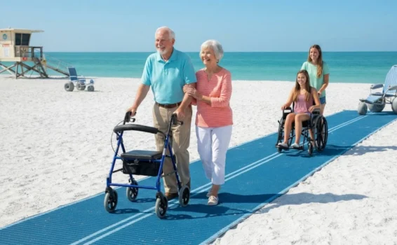 Two smiling seniors walk arm-in-arm on a blue mat at the beach, with a young woman pushing another young woman in a wheelchair behind them. The sea and sky are bright and clear in the background.