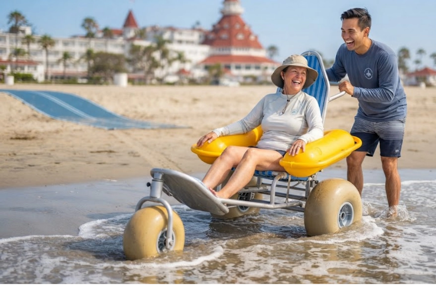 A smiling woman in a large beach wheelchair is pushed by a man along the shoreline, with a sunny beach, ocean, and a hotel in the background.