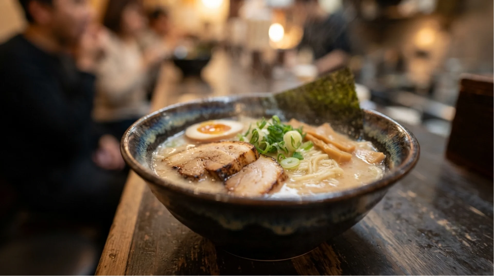 A bowl of ramen with sliced pork, half a boiled egg, green onions, bamboo shoots, and seaweed sits on a wooden counter in a restaurant, with blurred people in the background.