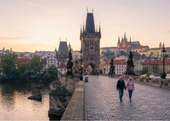 Two people walk along the historic Charles Bridge in Prague at sunrise, with city buildings, statues, and the Prague Castle visible in the background.