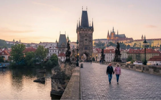 Two people walk along the historic Charles Bridge in Prague at sunrise, with city buildings, statues, and the Prague Castle visible in the background.