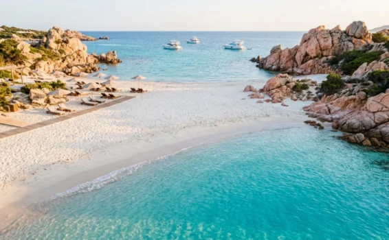Aerial view of a tropical beach with white sand, clear turquoise water, rocky cliffs, lounge chairs, and several yachts anchored in the distance.