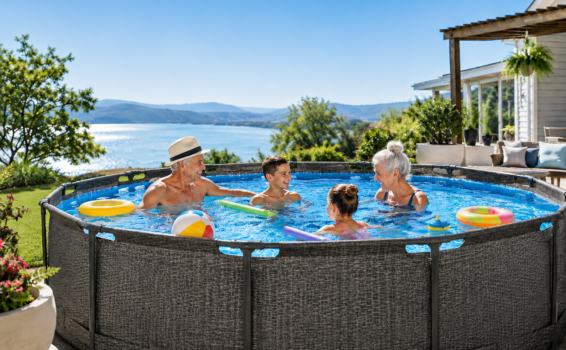 Two smiling adults and two children play with pool toys in a round above-ground pool, overlooking a scenic lake and hills under a clear blue sky. A house and garden are visible in the background.