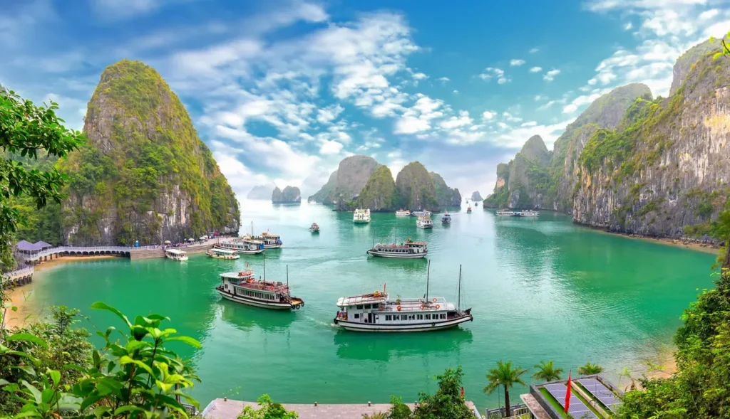 Boats float on vibrant green water surrounded by tall, rocky islands covered in greenery under a bright, partly cloudy sky.