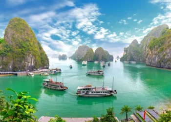 Boats float on vibrant green water surrounded by tall, rocky islands covered in greenery under a bright, partly cloudy sky.