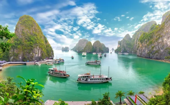 Boats float on vibrant green water surrounded by tall, rocky islands covered in greenery under a bright, partly cloudy sky.