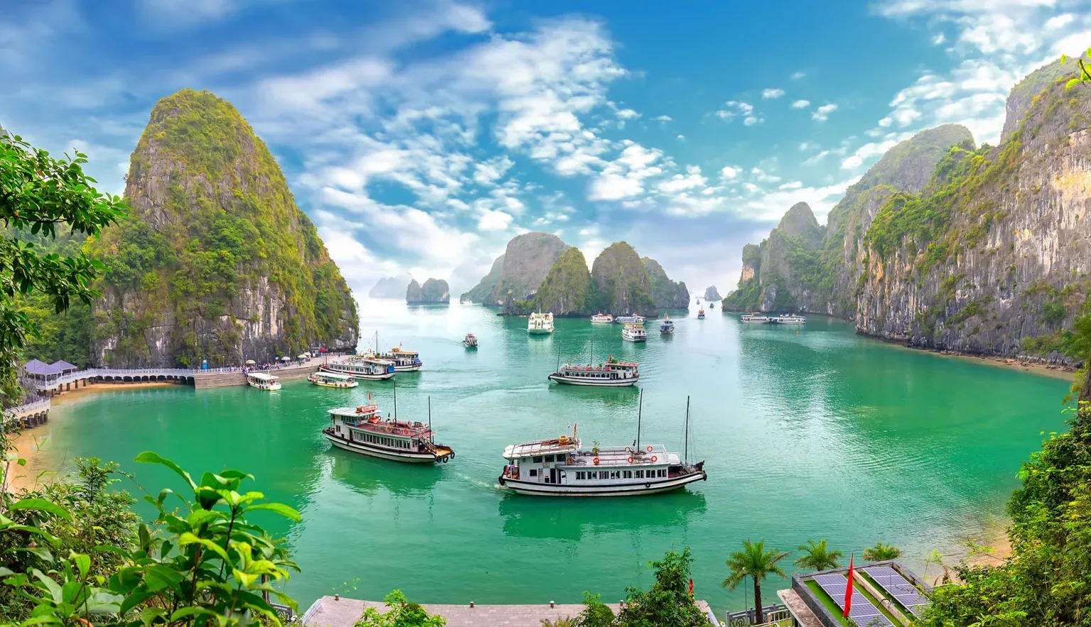 Boats float on vibrant green water surrounded by tall, rocky islands covered in greenery under a bright, partly cloudy sky.