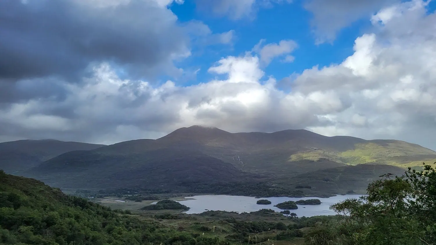 A scenic view of a lake with small islands, surrounded by green hills and mountains under a partly cloudy sky with patches of blue.