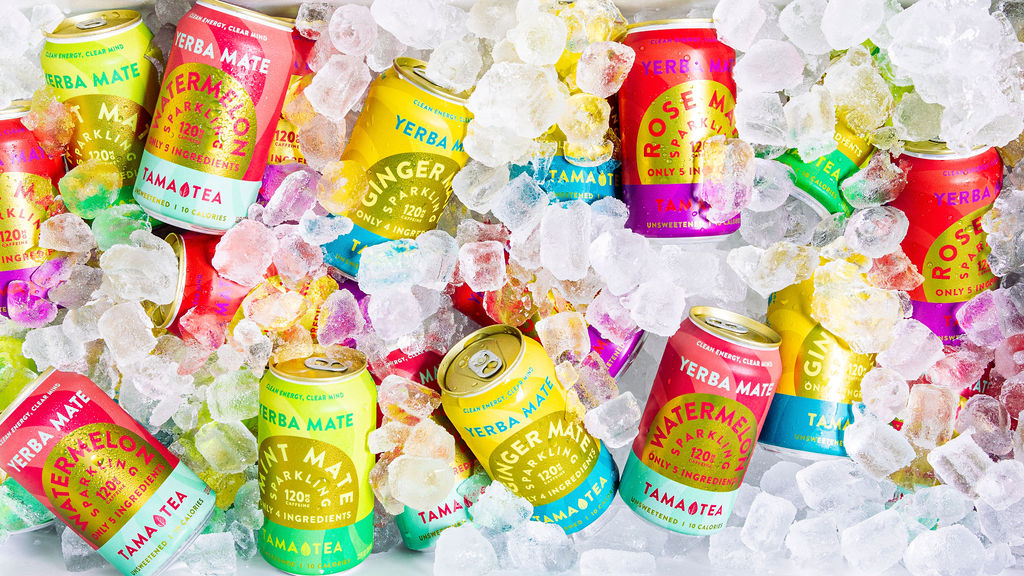 Assorted colorful cans of Yerba Mate Tamatea drinks are scattered among ice cubes, with flavors like ginger, watermelon, and rose visible on the labels.